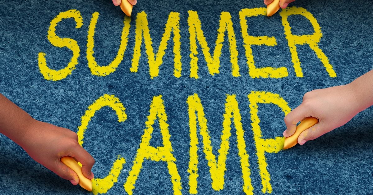 four hands holding yellow chalk write summer camp in yellow letters on the ground